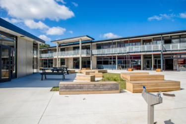 Koru school shown on a sunny day, a courtyard is shown in the foreground with two rows of classrooms in the background