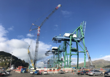 Industrial port cranes are shown on a sunny day with machinery and shipping containers in the foreground.